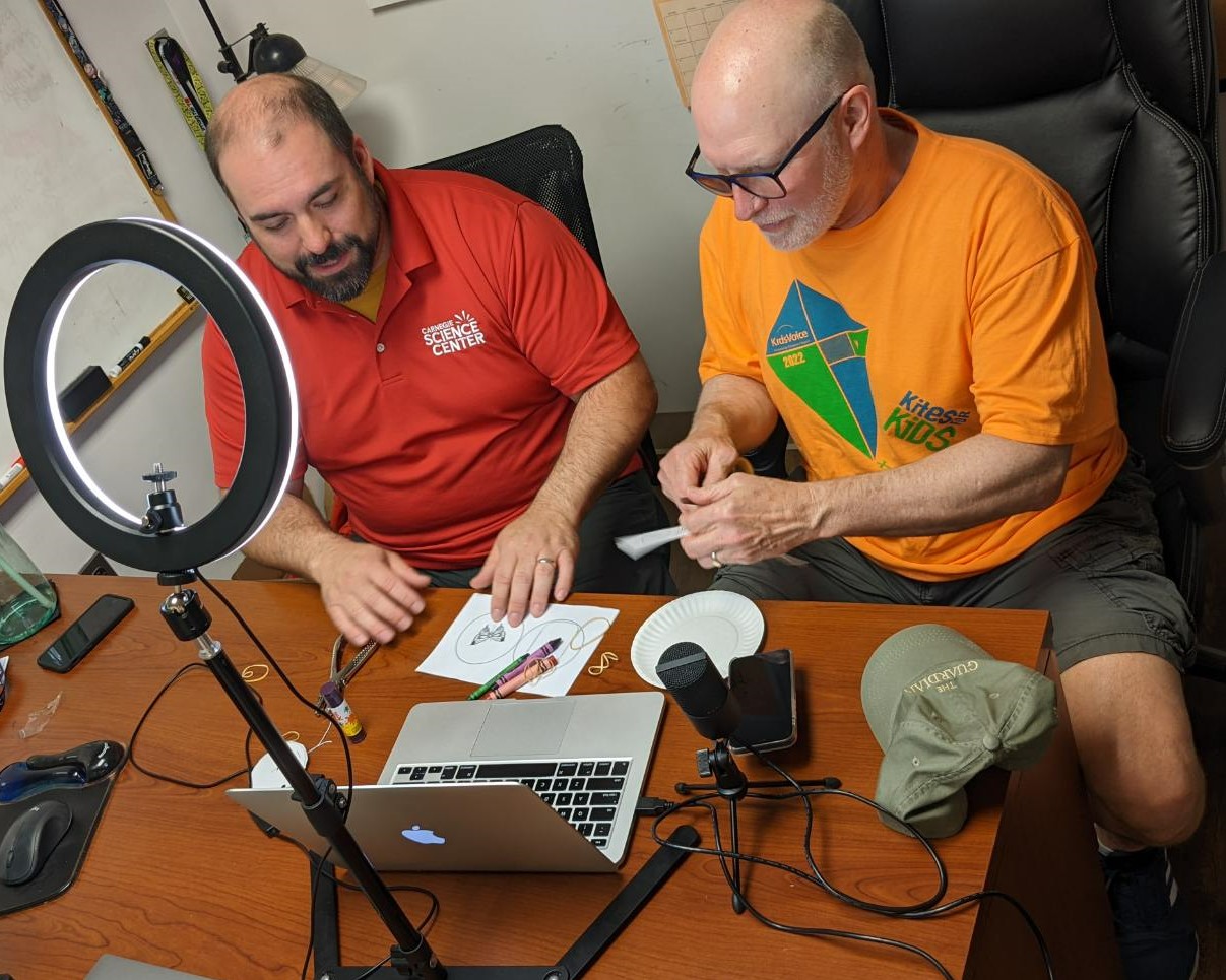 A ring light puts a spotlight on two men sitting at a desk in front of a laptop. One man is cutting paper.