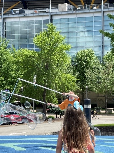 A man in an orange shirt uses large rods to create a giant bubbles
