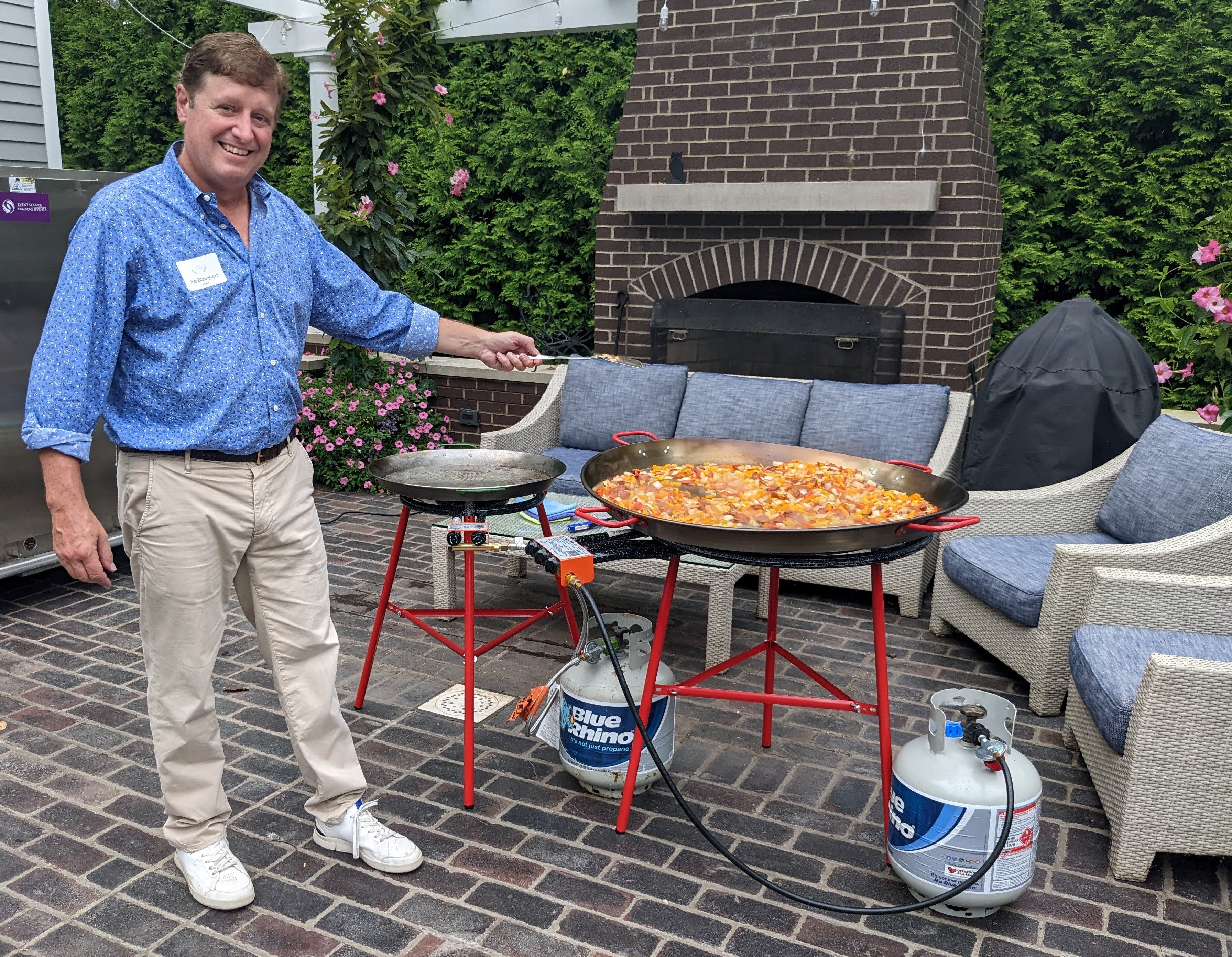 A man stands beside a large pan of paella in an outdoor kitchen
