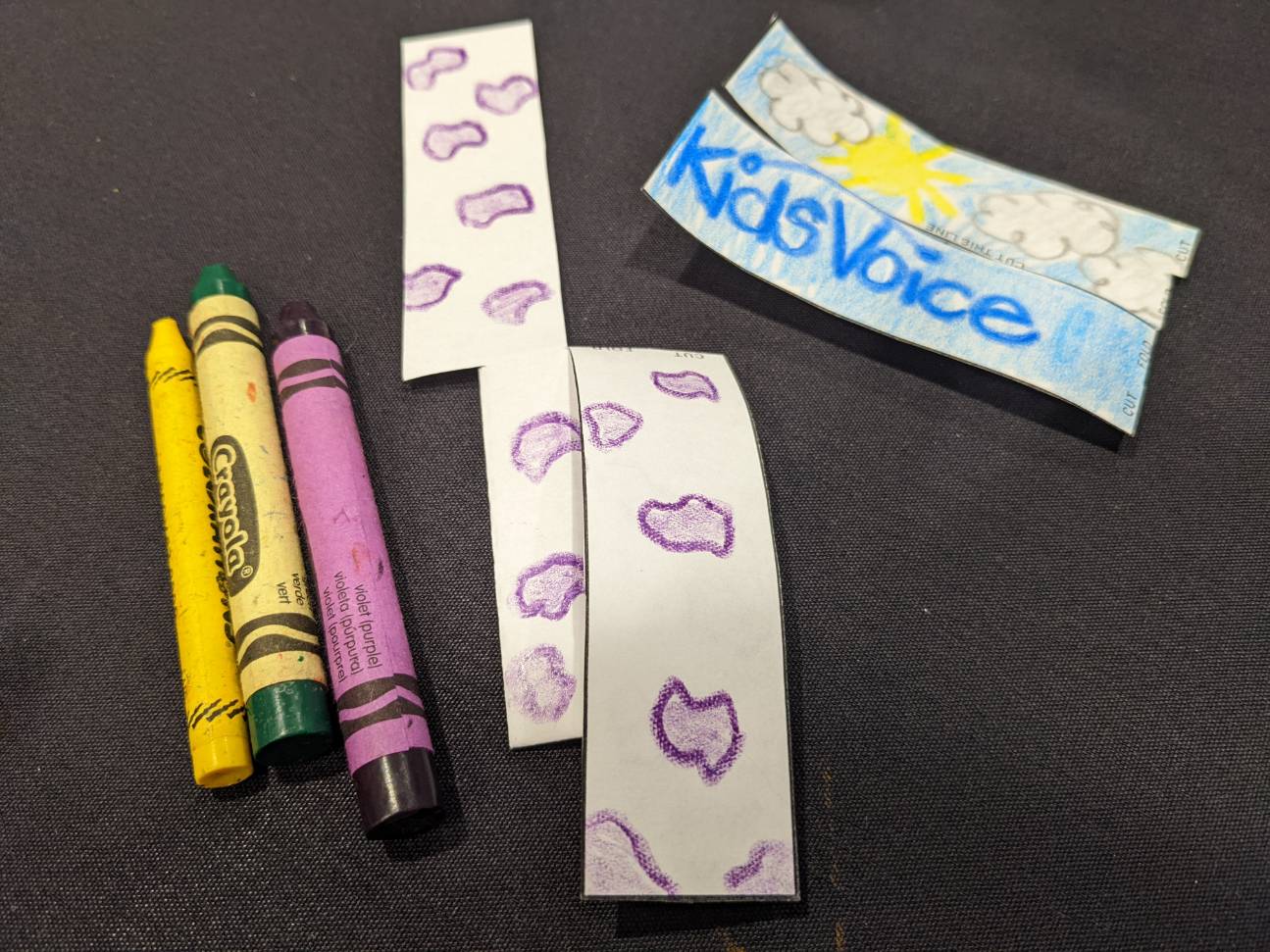 Three crayons lay next to two strips of paper, one decorated with purple spots and the other with clouds, the sun, and the word KidsVoice 