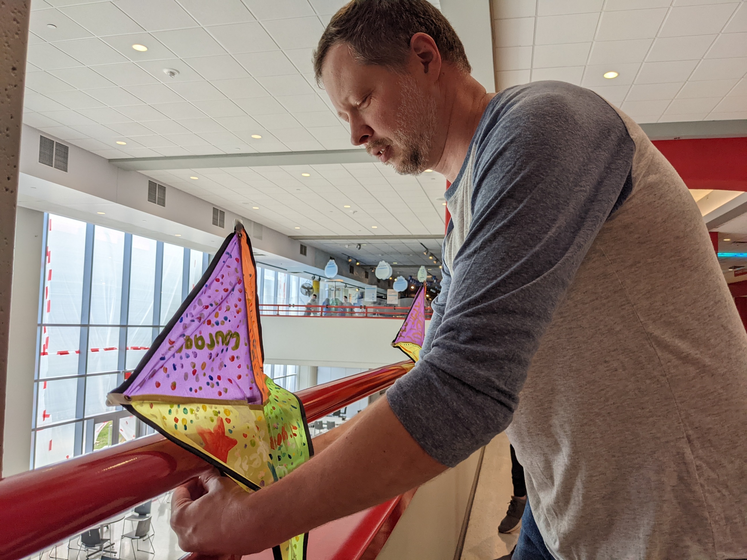 A man in a blue and grey shirt hangs a decorated kite in the Carnegie Science Center lobby